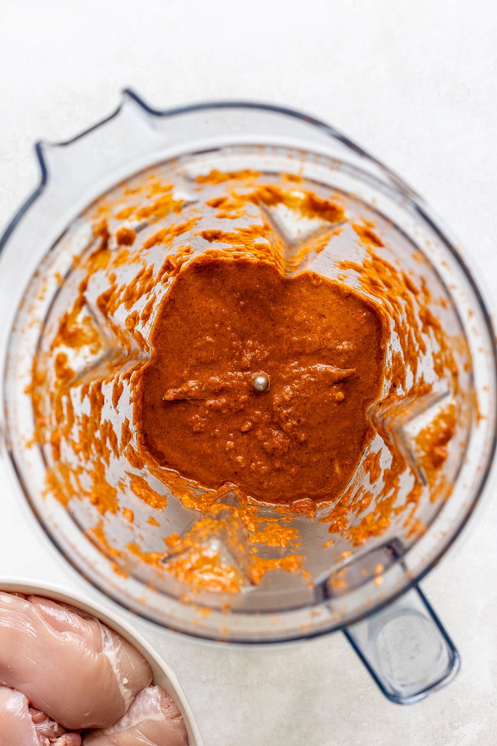 Overhead view of a blender containing an orange-red sauce mixture, with a bowl of raw chicken pieces partially visible in the corner.