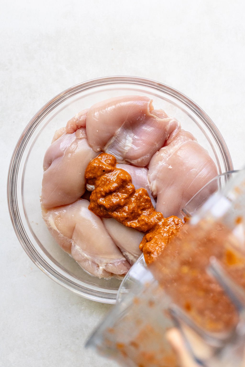 Raw chicken thighs in a glass bowl with a reddish-brown marinade being poured over them from a food processor.