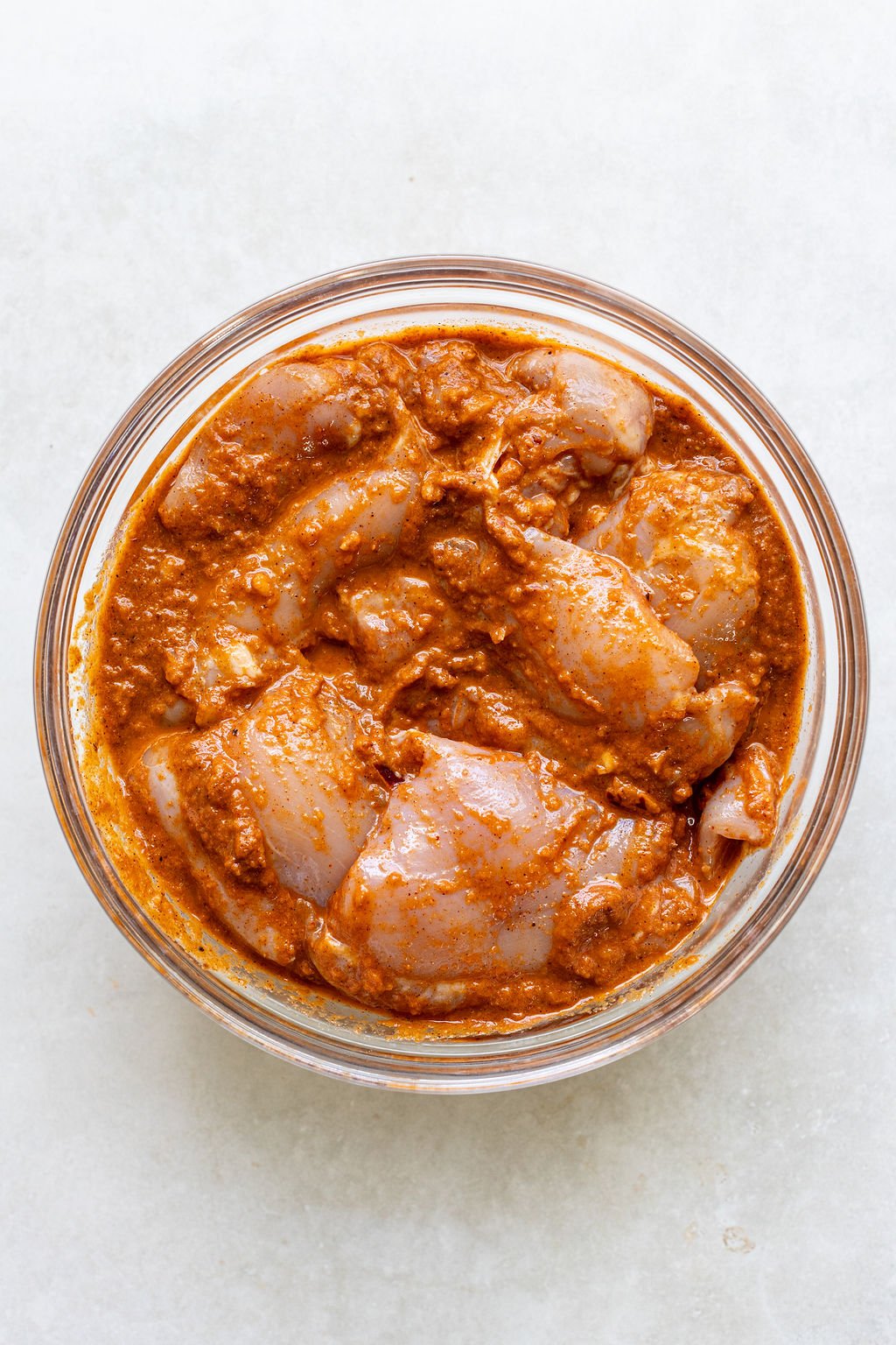 Raw chicken pieces coated in a reddish-orange marinade in a clear glass bowl on a light surface.
