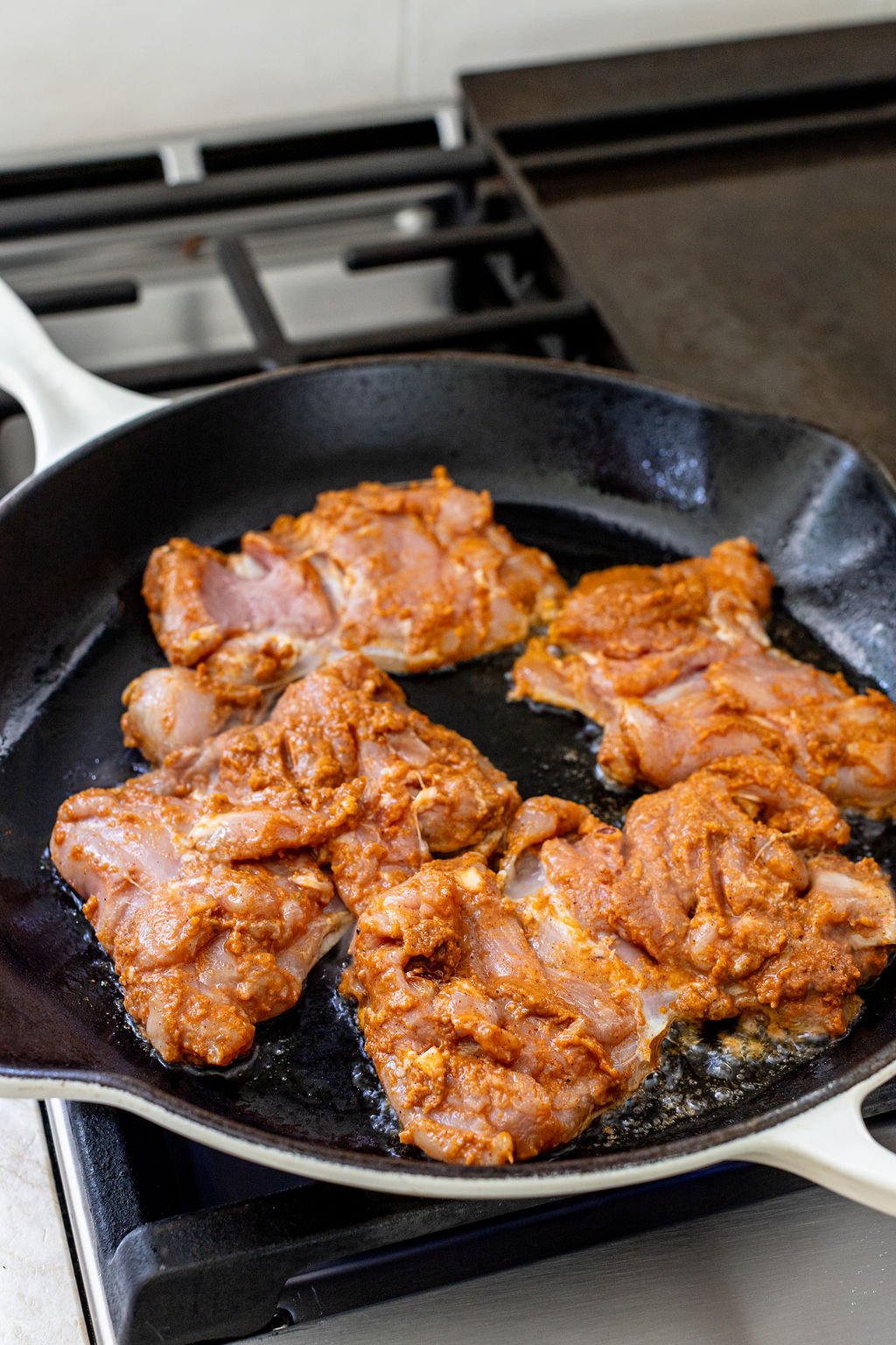 Four pieces of marinated chicken are being cooked in a black cast iron skillet on a stovetop.