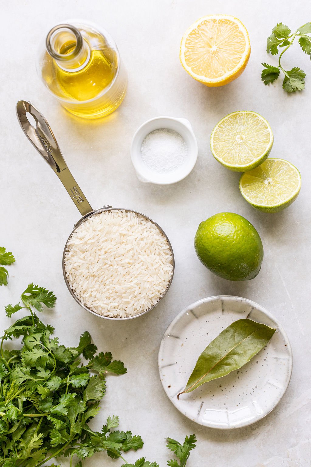 A measuring cup of uncooked white rice, a halved lemon, a halved lime, a whole lime, a small bowl of salt, a bottle of oil, fresh cilantro, and a plate with bay leaves on a white surface.