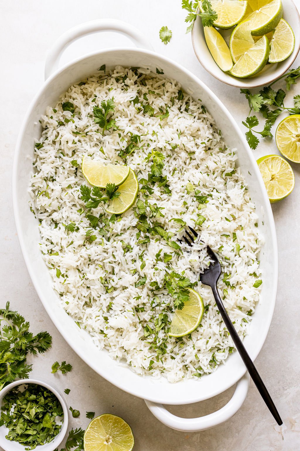 White rice garnished with chopped cilantro and lime wedges in a white oval dish, with a black fork and extra lime and cilantro on the side.