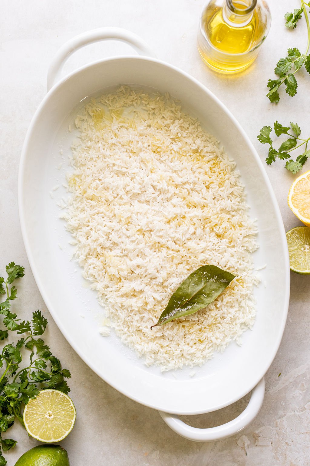 An oval white dish with uncooked rice, a bay leaf, and oil.