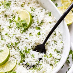 A black fork rests in a white dish filled with cilantro lime rice, garnished with lime wedges and fresh cilantro.