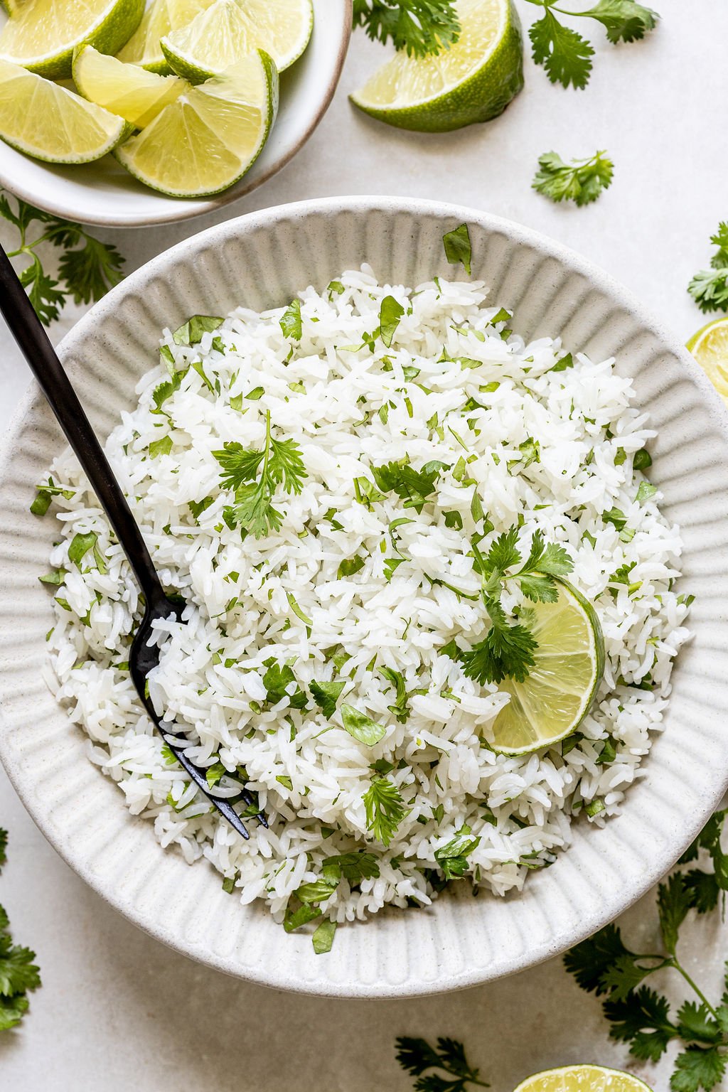 A bowl of white rice garnished with chopped cilantro and a lime wedge, with a black fork in the bowl and lime slices and cilantro scattered around.