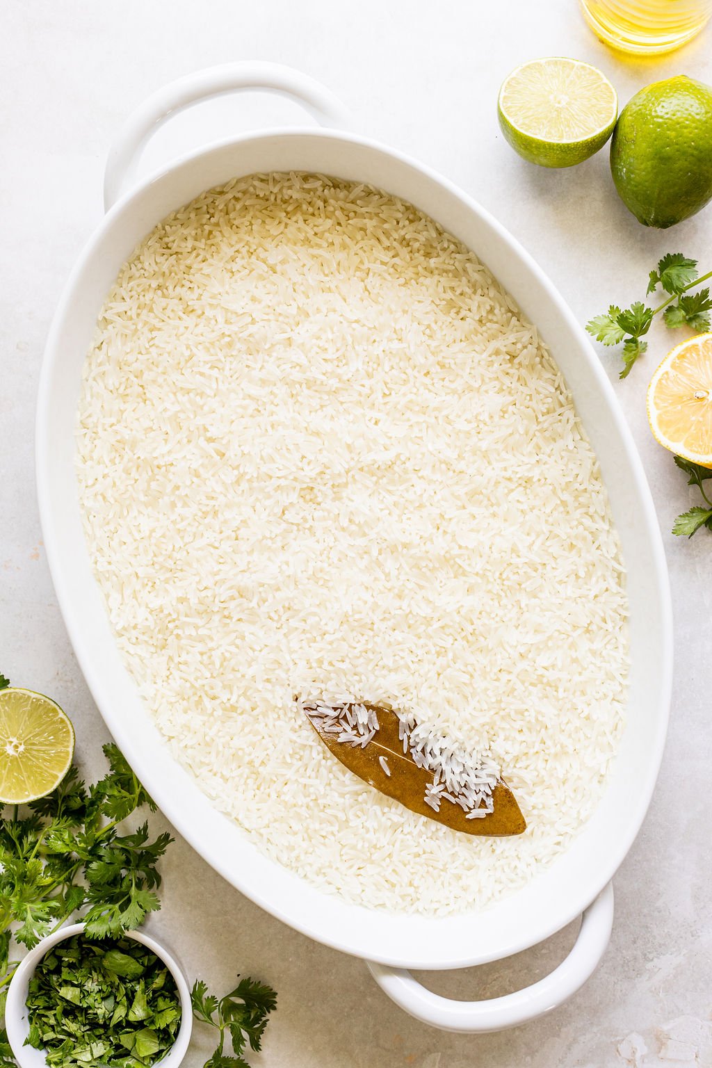 Oval white dish filled with uncooked white rice, topped with a bay leaf, surrounded by lime halves, fresh cilantro, and a bottle of oil on a light surface.