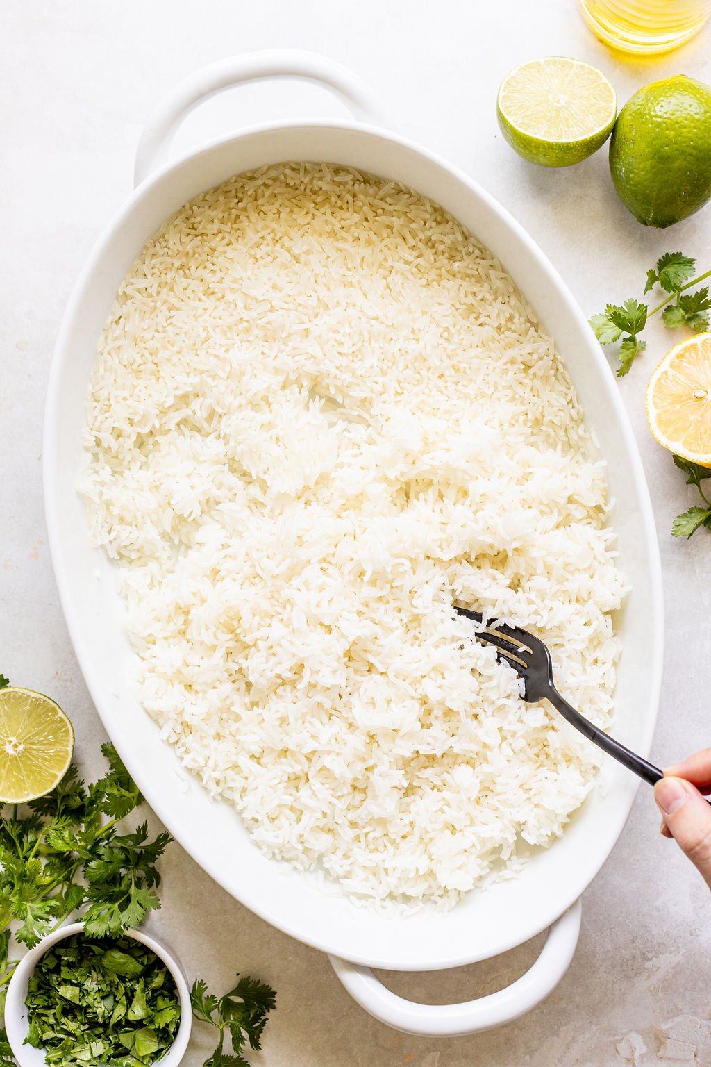 A hand uses a fork to fluff white rice in an oval dish, surrounded by halved limes, fresh cilantro, and a small bowl of chopped herbs on a light surface.