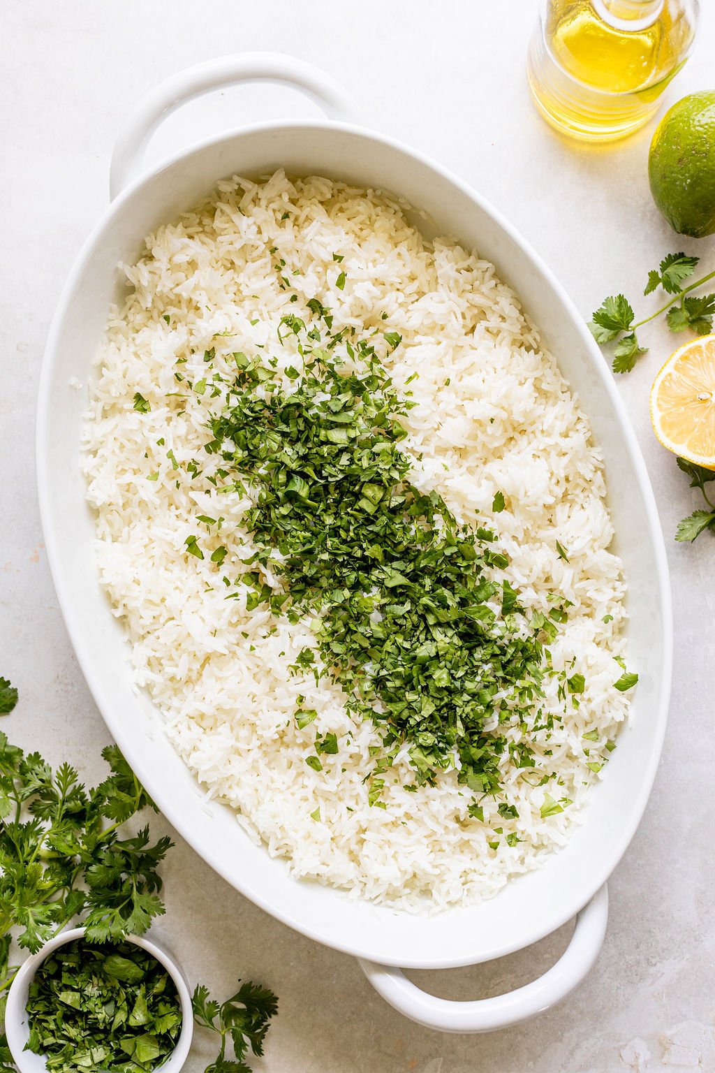 A white oval dish filled with cooked white rice, topped with a line of chopped cilantro. Lemon, lime, olive oil, and extra cilantro are arranged nearby.