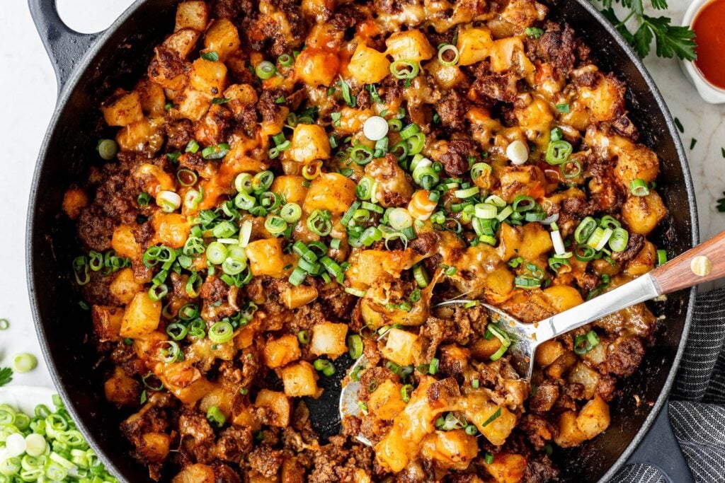 A skillet filled with cooked ground beef, diced potatoes, melted cheese, and chopped green onions, with a serving spoon partially scooping the mixture.