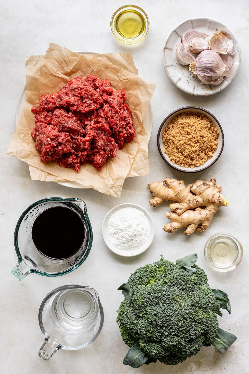 Ingredients arranged on a white surface, including ground beef, broccoli, fresh ginger, garlic, brown sugar, flour, soy sauce, vegetable oil, and water.