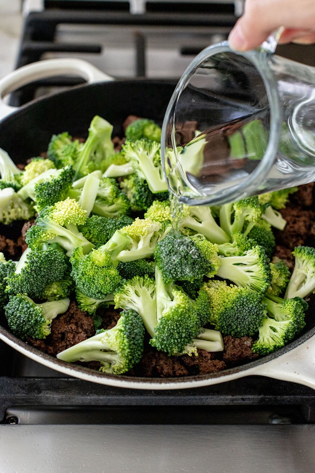 A hand pours water from a glass measuring cup over broccoli and ground meat cooking in a skillet on a stovetop.