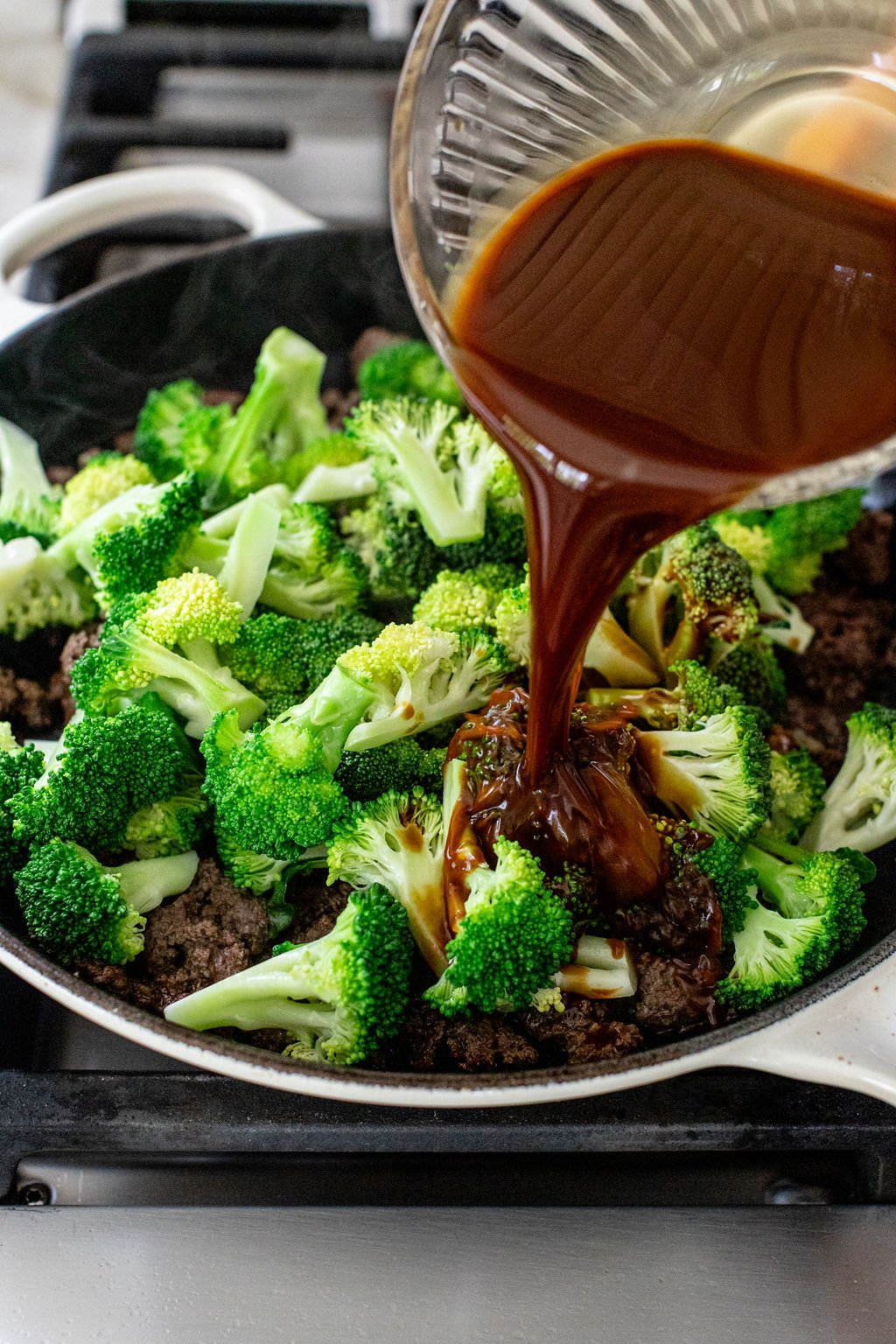 Broccoli and ground beef in a skillet topped with brown sauce being poured from a glass container.