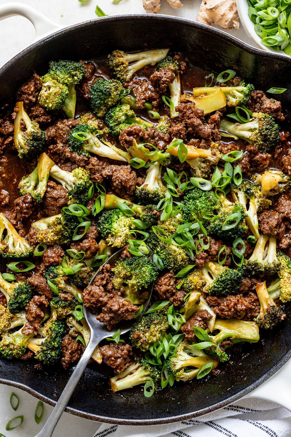 A skillet filled with cooked ground beef, broccoli florets, and sliced green onions, with a serving spoon partially in the dish.