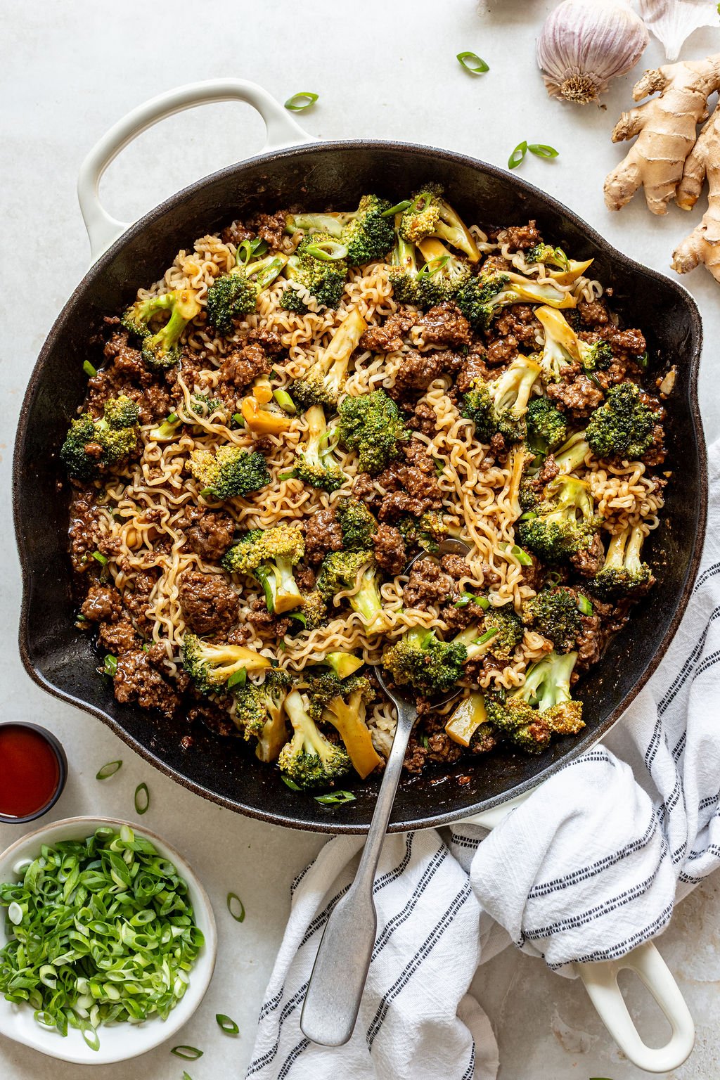 A skillet filled with cooked noodles, ground beef, and broccoli, garnished with green onions, with a serving spoon and fresh ingredients nearby.