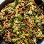 A skillet filled with cooked ramen noodles, ground beef, broccoli, and green onions, with a spoon on the side.