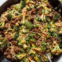 A skillet filled with cooked ramen noodles, ground beef, broccoli, and green onions, with a spoon on the side.