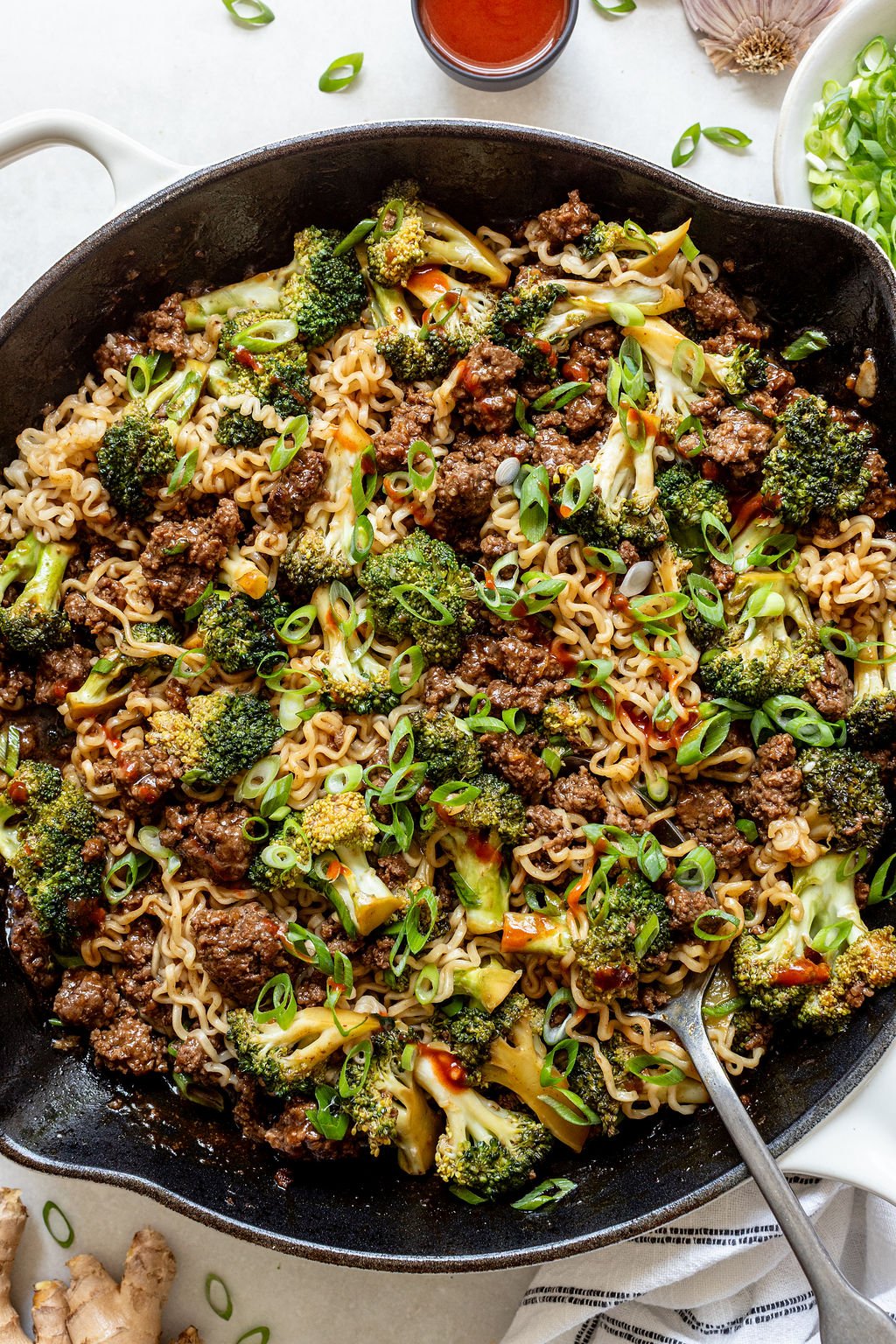 A skillet filled with cooked ramen noodles, ground beef, broccoli, and green onions, with a spoon on the side.