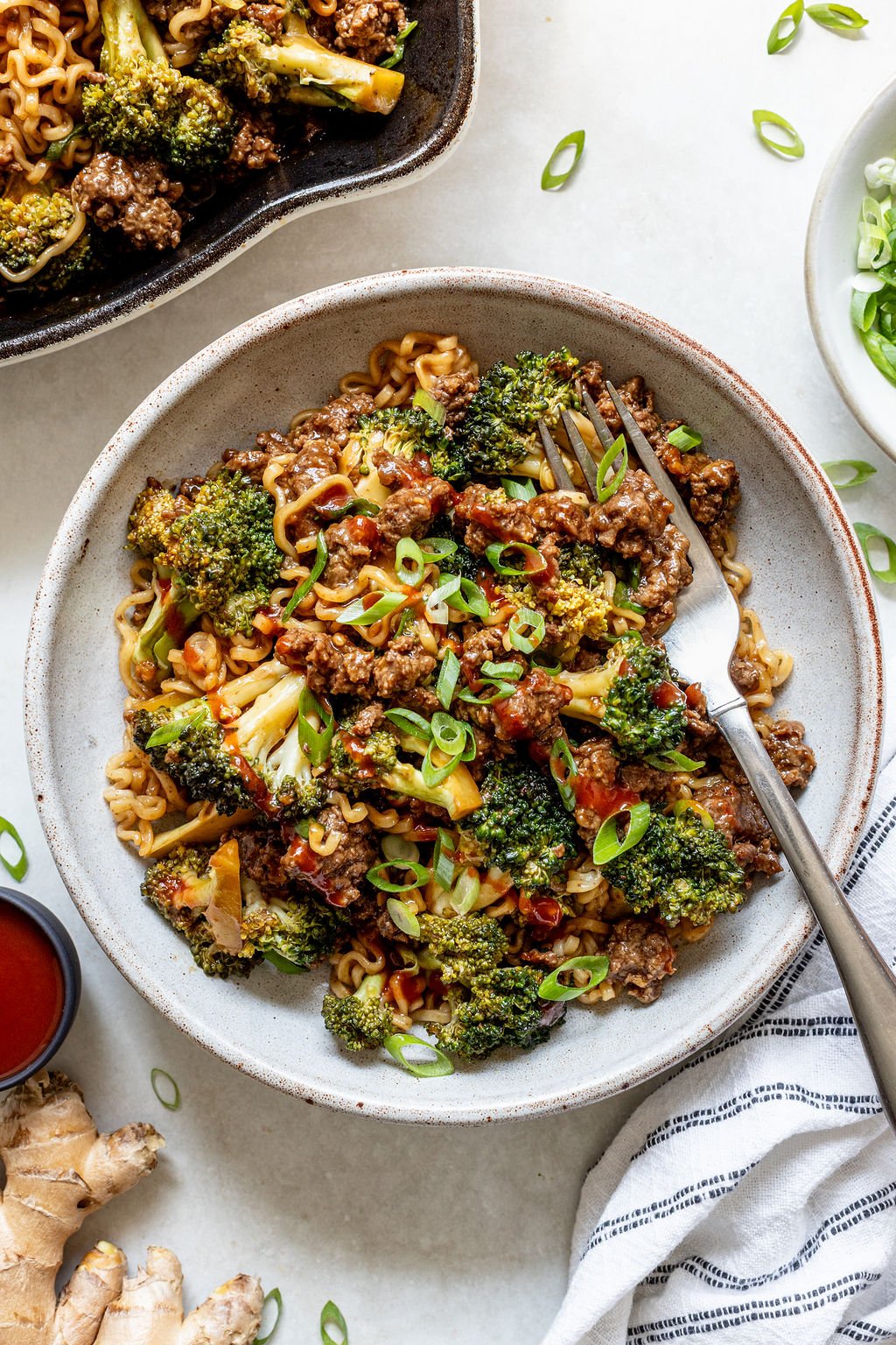 A bowl of stir-fried noodles with ground beef, broccoli, and scallions, served with a fork. A plate of the same dish, fresh ginger, and a small bowl of sauce are nearby.