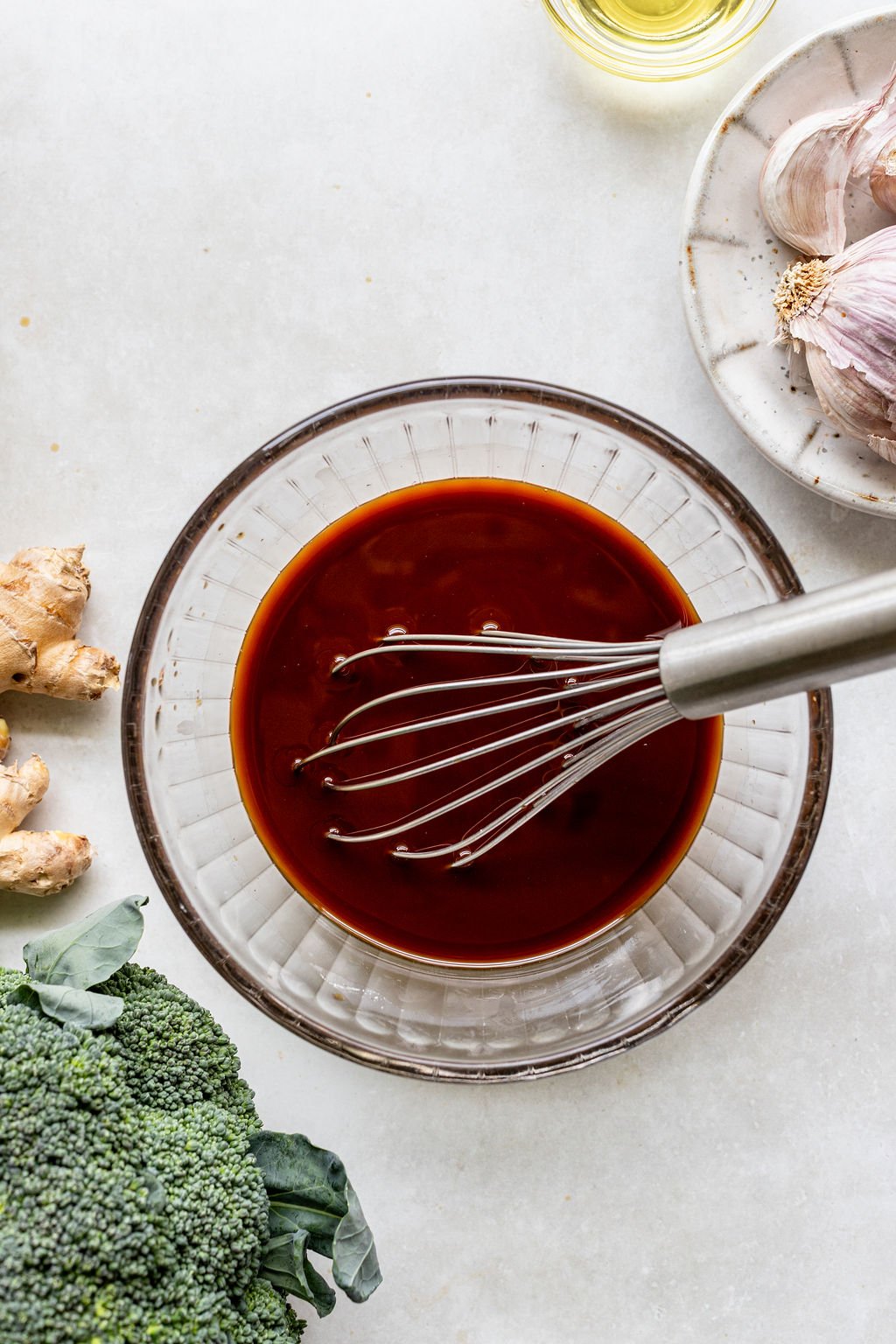 A glass bowl with dark sauce and a whisk, surrounded by broccoli, ginger, garlic, and a small dish of oil on a light countertop.