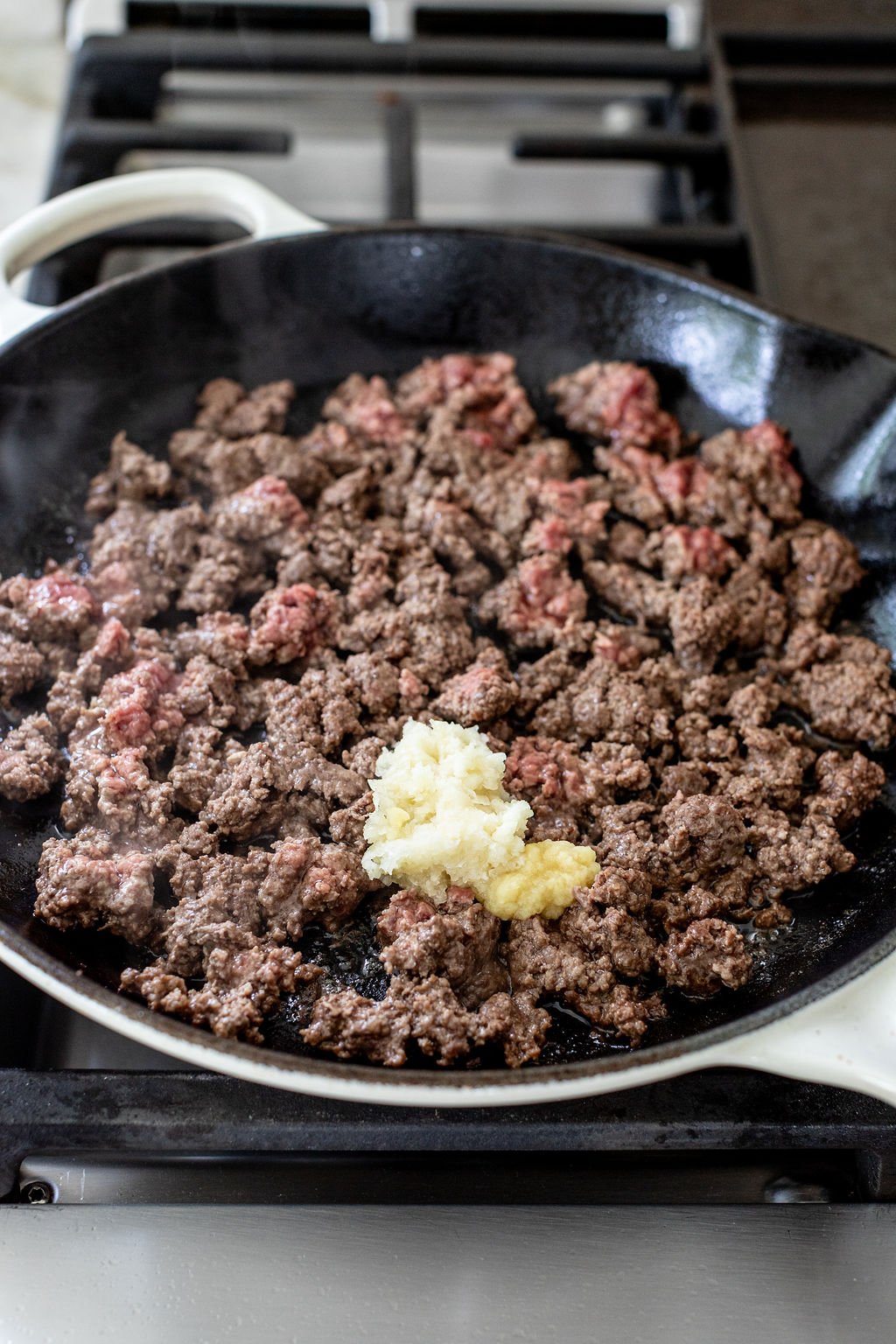 Ground beef cooking in a skillet on a stovetop with a mound of minced garlic added to the center.