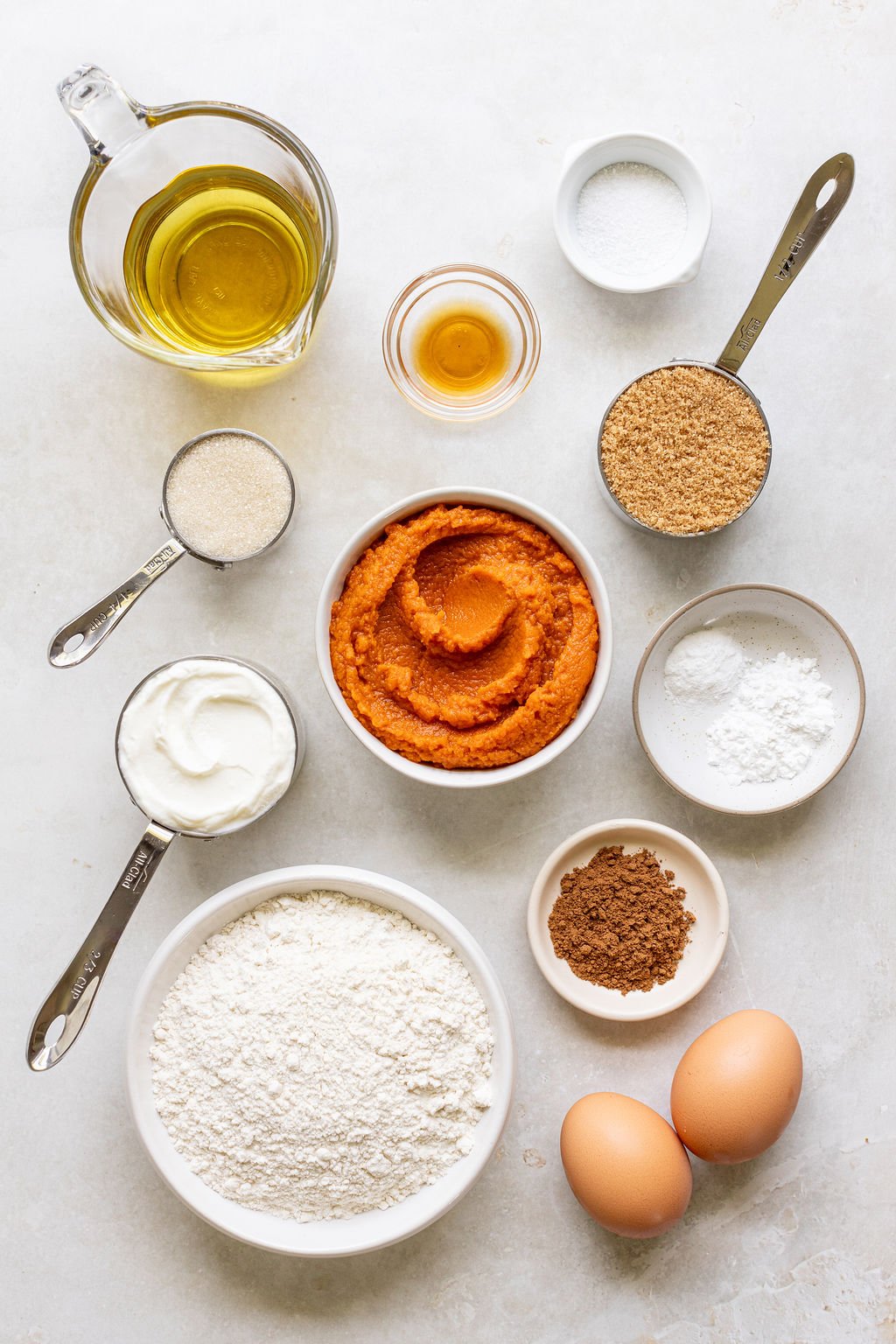 Overhead view of measured baking ingredients including flour, sugar, oil, eggs, pumpkin puree, yogurt, spices, baking powder, baking soda, and vanilla on a white surface.