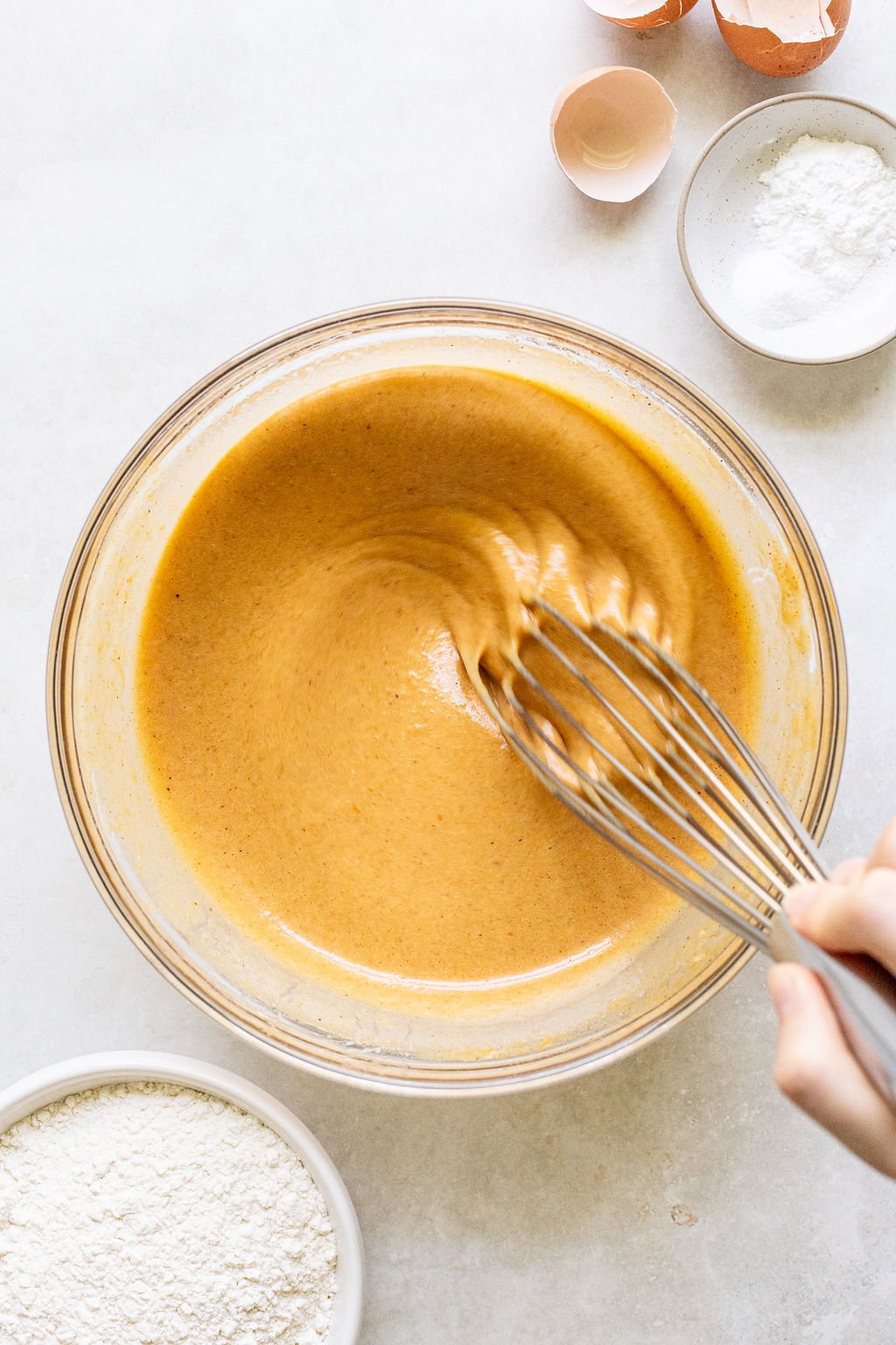 A hand whisking a bowl of light brown batter, with bowls of flour, baking powder, and eggshells on a white surface nearby.