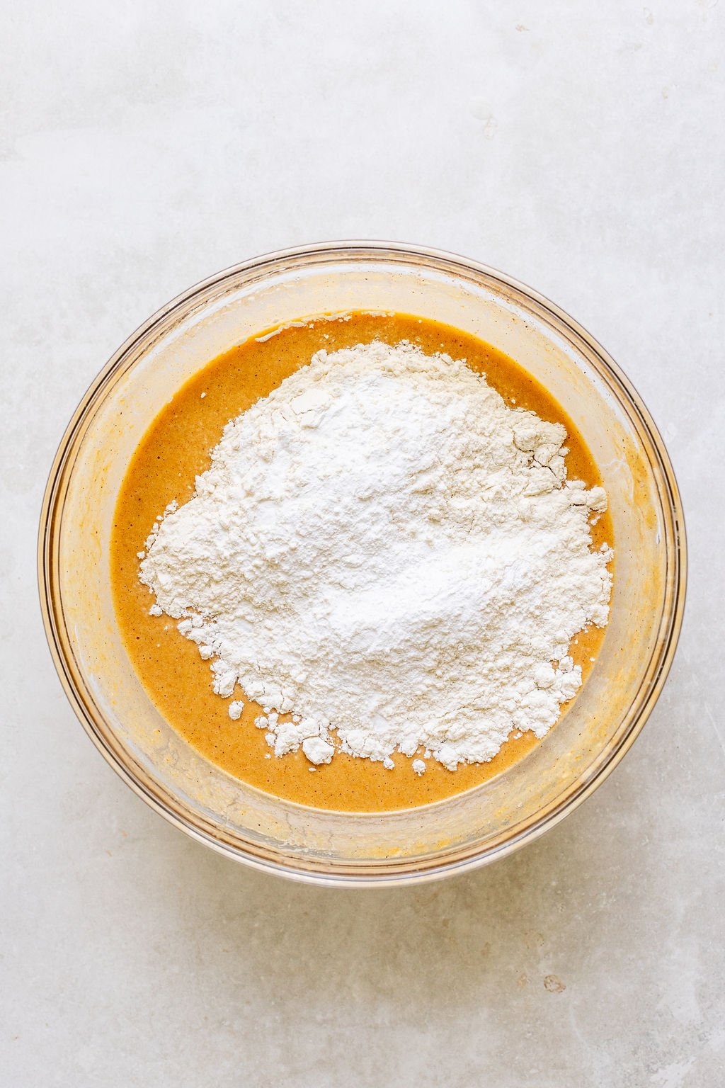 A glass bowl with orange-brown batter and a mound of white flour on top, sitting on a light-colored surface.