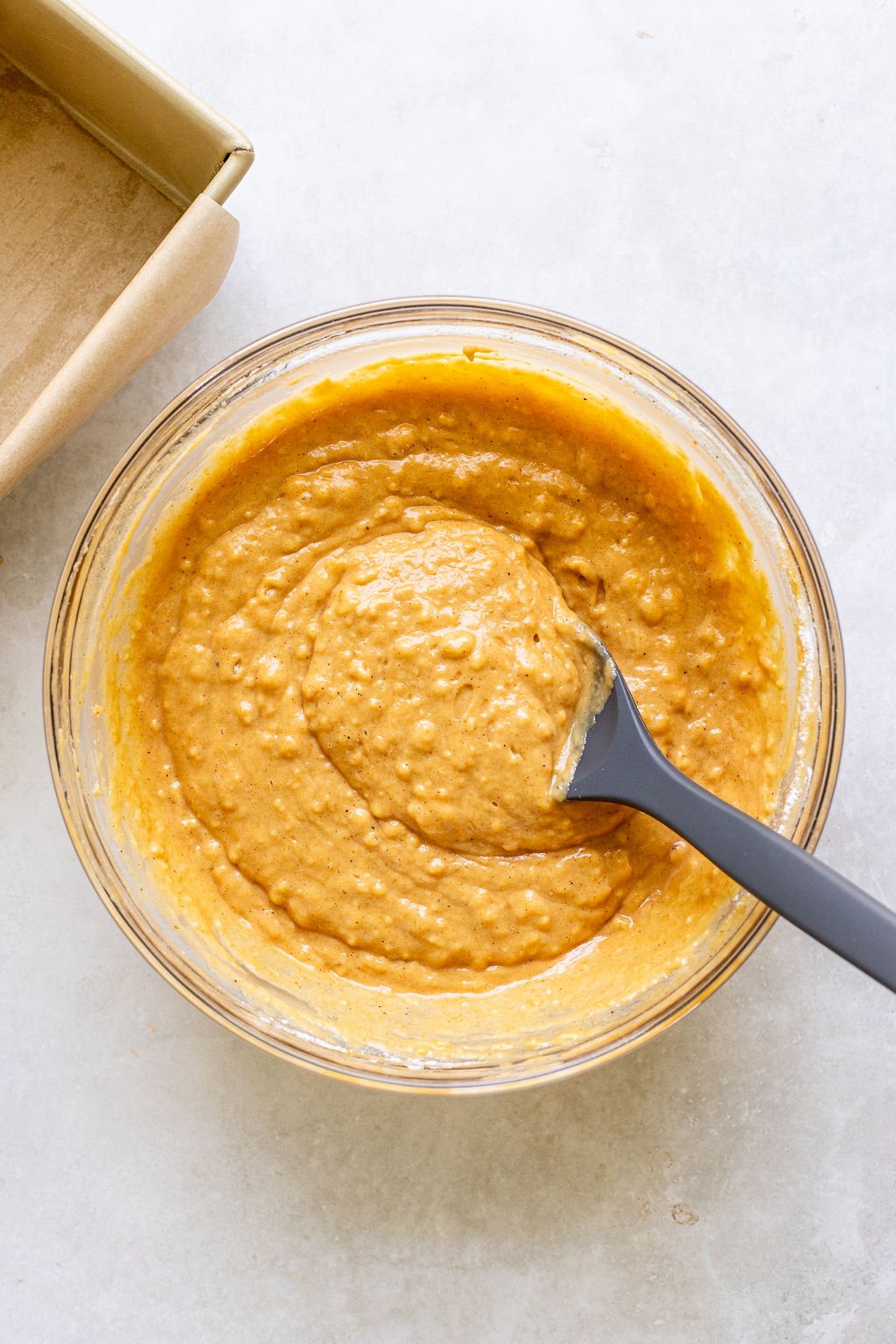 A glass bowl filled with thick, orange batter and a black spatula on a light countertop, next to a parchment-lined baking pan.