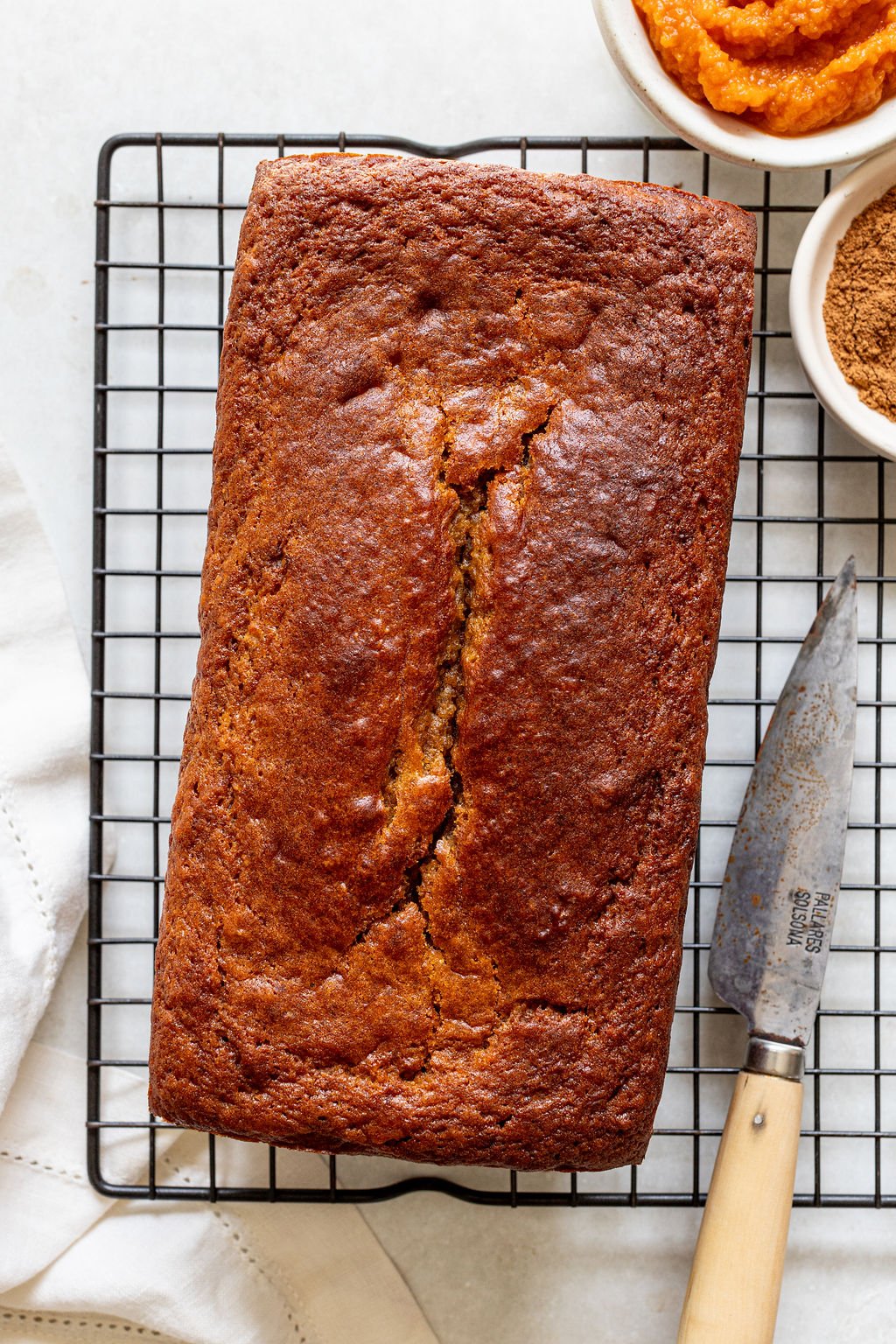 A loaf of pumpkin bread sits on a black cooling rack next to a knife and small bowls of pumpkin puree and ground cinnamon.