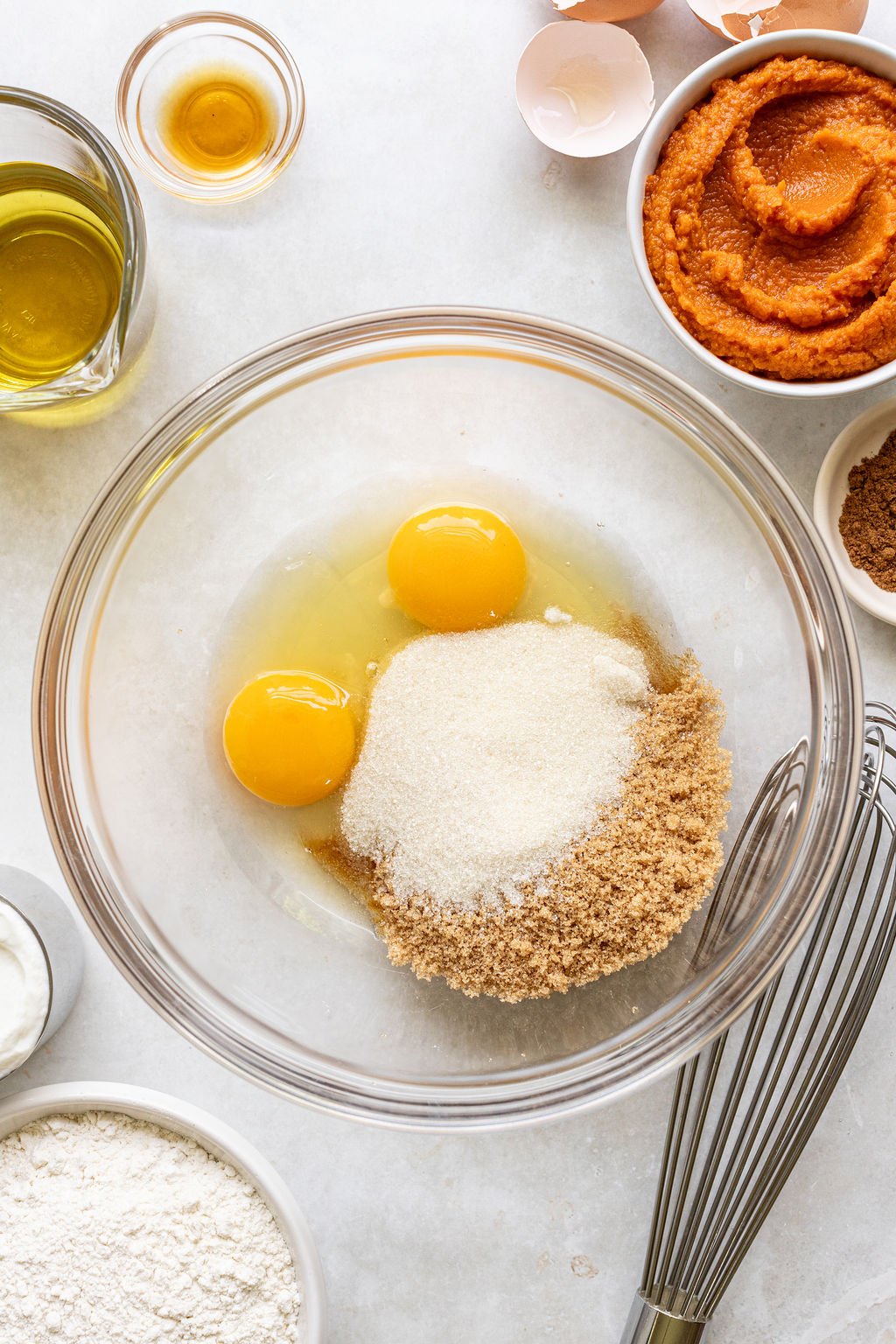 A glass bowl with eggs, brown sugar, and white sugar, surrounded by flour, pumpkin puree, oil, spices, a whisk, and cracked eggshells on a white surface.