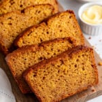 Several slices of pumpkin bread are arranged on a wooden board, with small bowls of butter and pumpkin puree in the background.