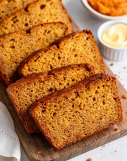 Several slices of pumpkin bread are arranged on a wooden board, with small bowls of butter and pumpkin puree in the background.
