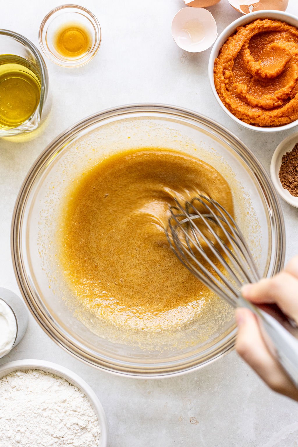 A hand whisks a brown mixture in a glass bowl surrounded by bowls of flour, pumpkin puree, oil, and other baking ingredients on a white surface.
