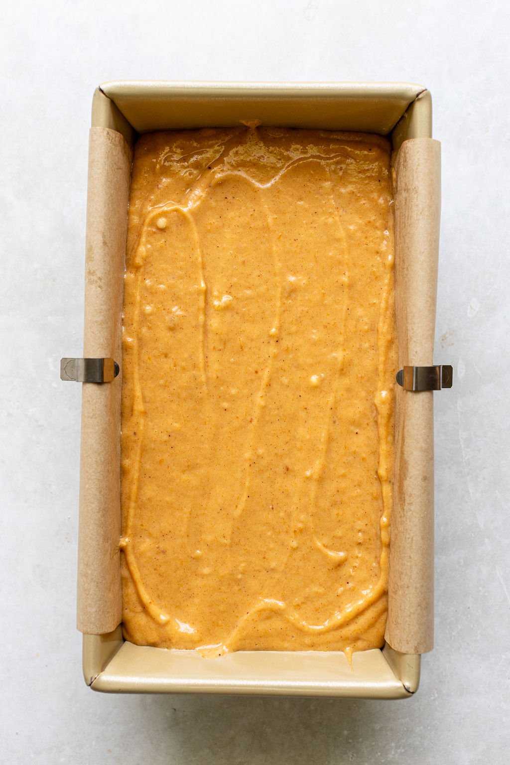 A loaf pan lined with parchment paper filled with smooth orange batter.