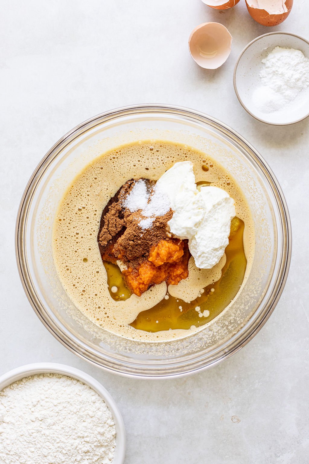 A glass bowl with eggs, brown sugar, pumpkin puree, yogurt, and spices, surrounded by bowls of flour, baking powder, and broken eggshells on a white surface.