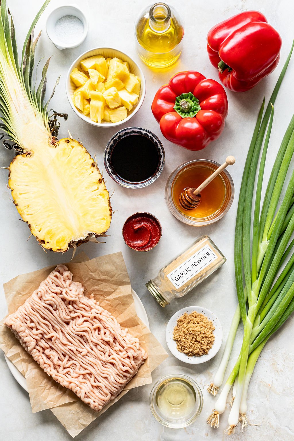 Overhead view of raw ground meat, pineapple, red bell peppers, green onions, brown sugar, honey, garlic powder, soy sauce, tomato paste, oil, and salt arranged on a white surface.