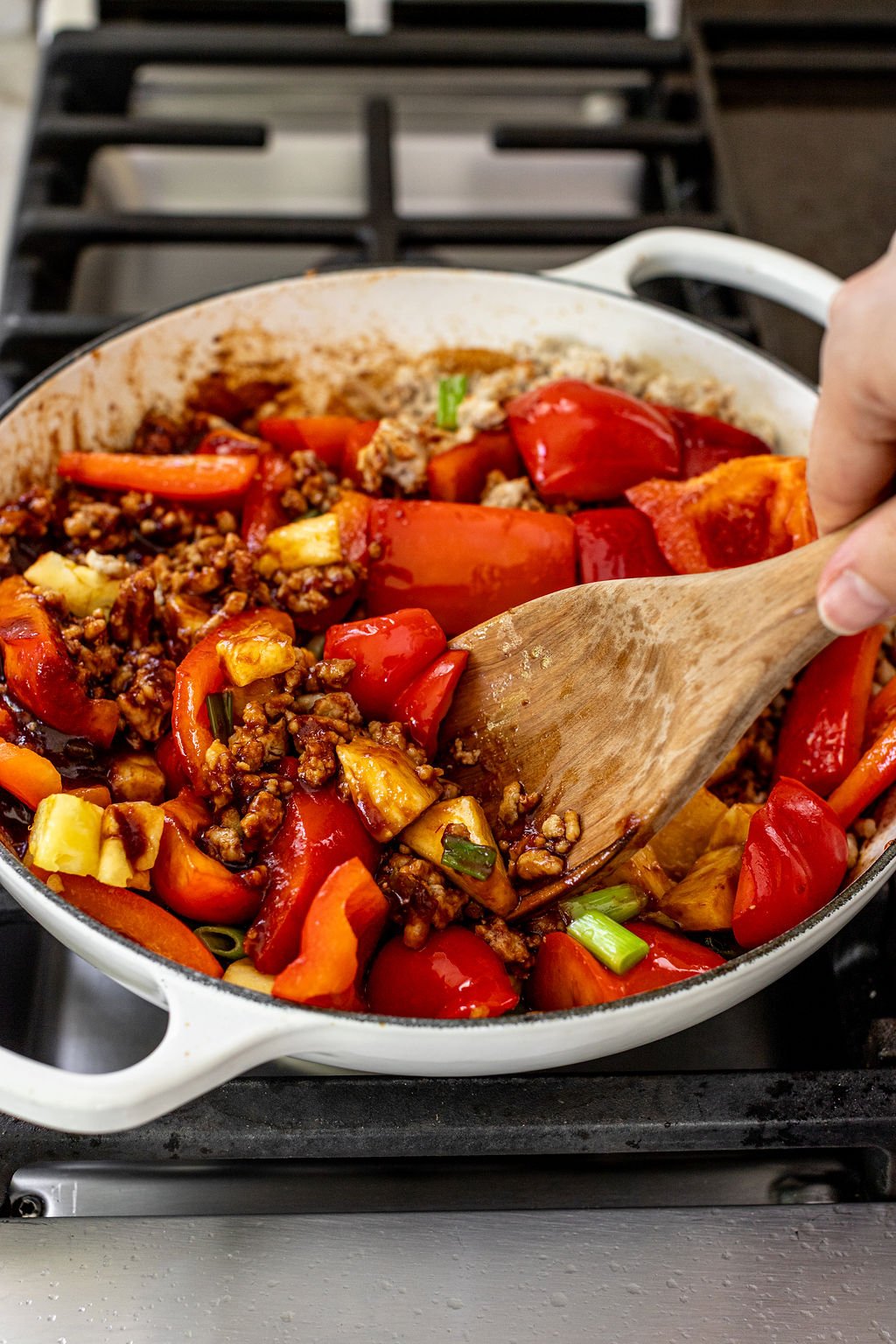 A hand stirs a mixture of red bell peppers, ground meat, and vegetables in a white skillet on a stovetop.