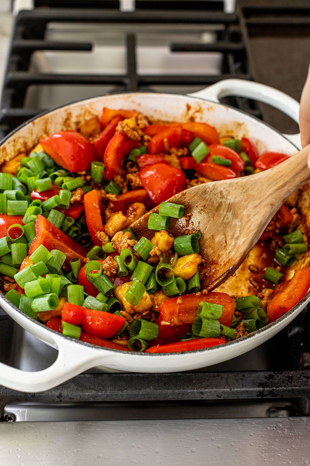 A wooden spoon stirs a pan of sautéed vegetables, including chopped green onions, red bell peppers, and ground meat, on a stovetop.