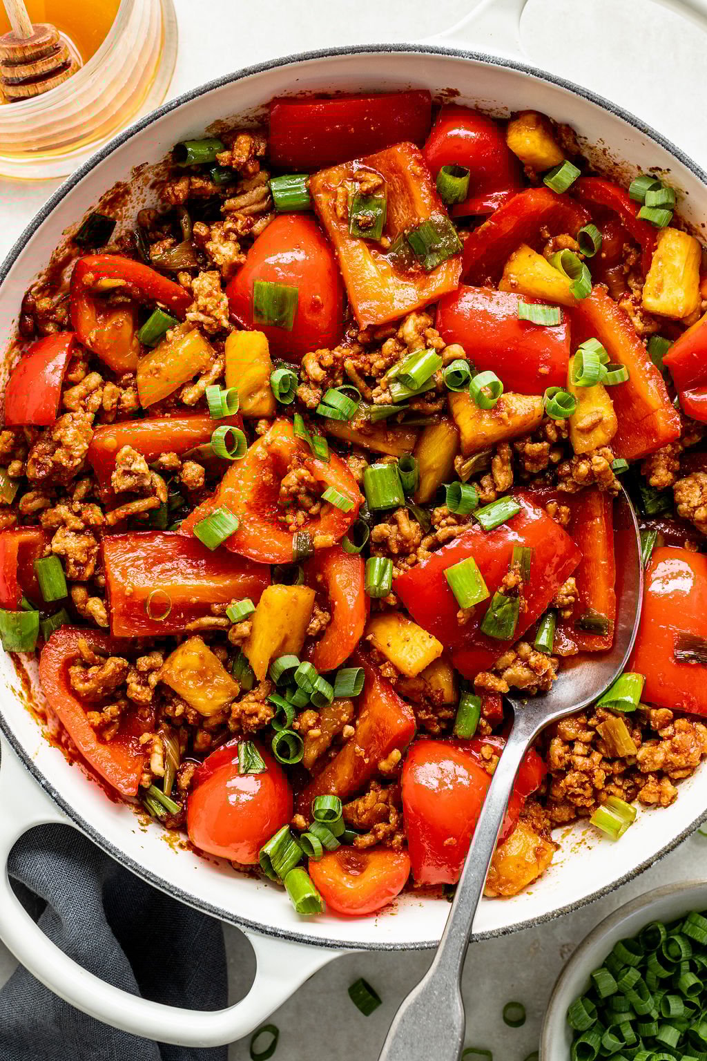 A skillet filled with stir-fried ground meat, red bell peppers, pineapple chunks, and chopped green onions, with a spoon resting in the dish.