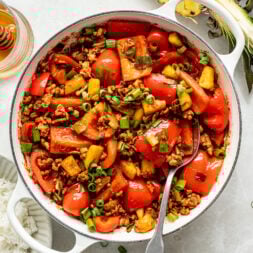 A white pot filled with a stir-fry of ground meat, red bell peppers, pineapple chunks, and green onions, with a spoon inside.