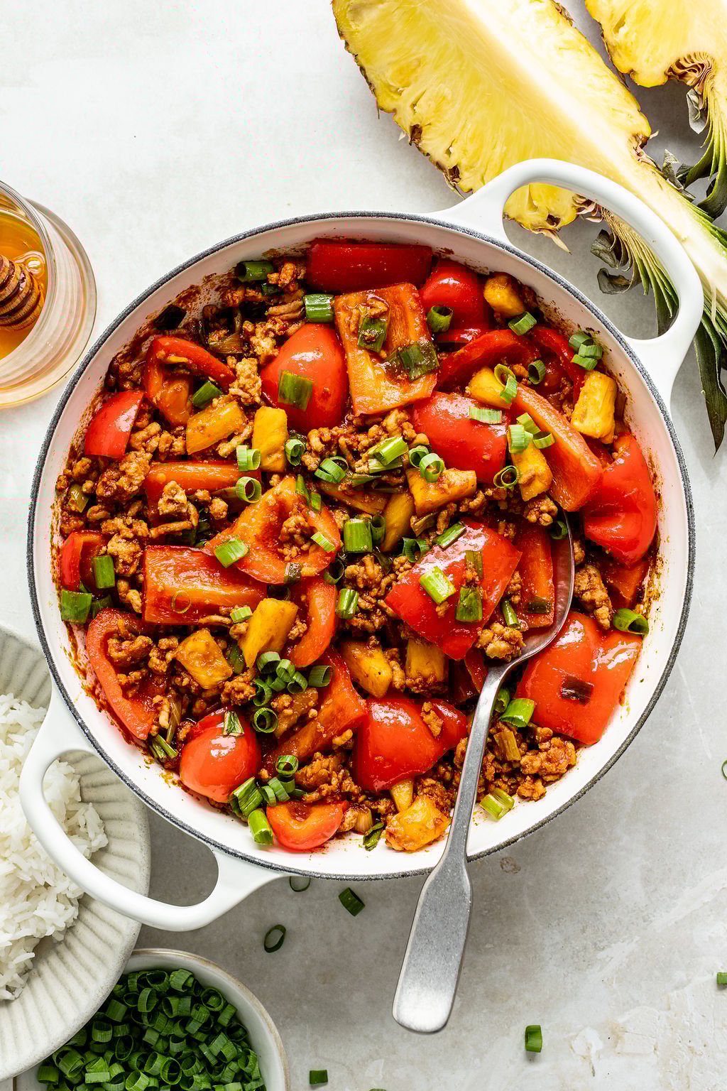 A white pot filled with a stir-fry of ground meat, red bell peppers, pineapple chunks, and green onions, with a spoon inside.