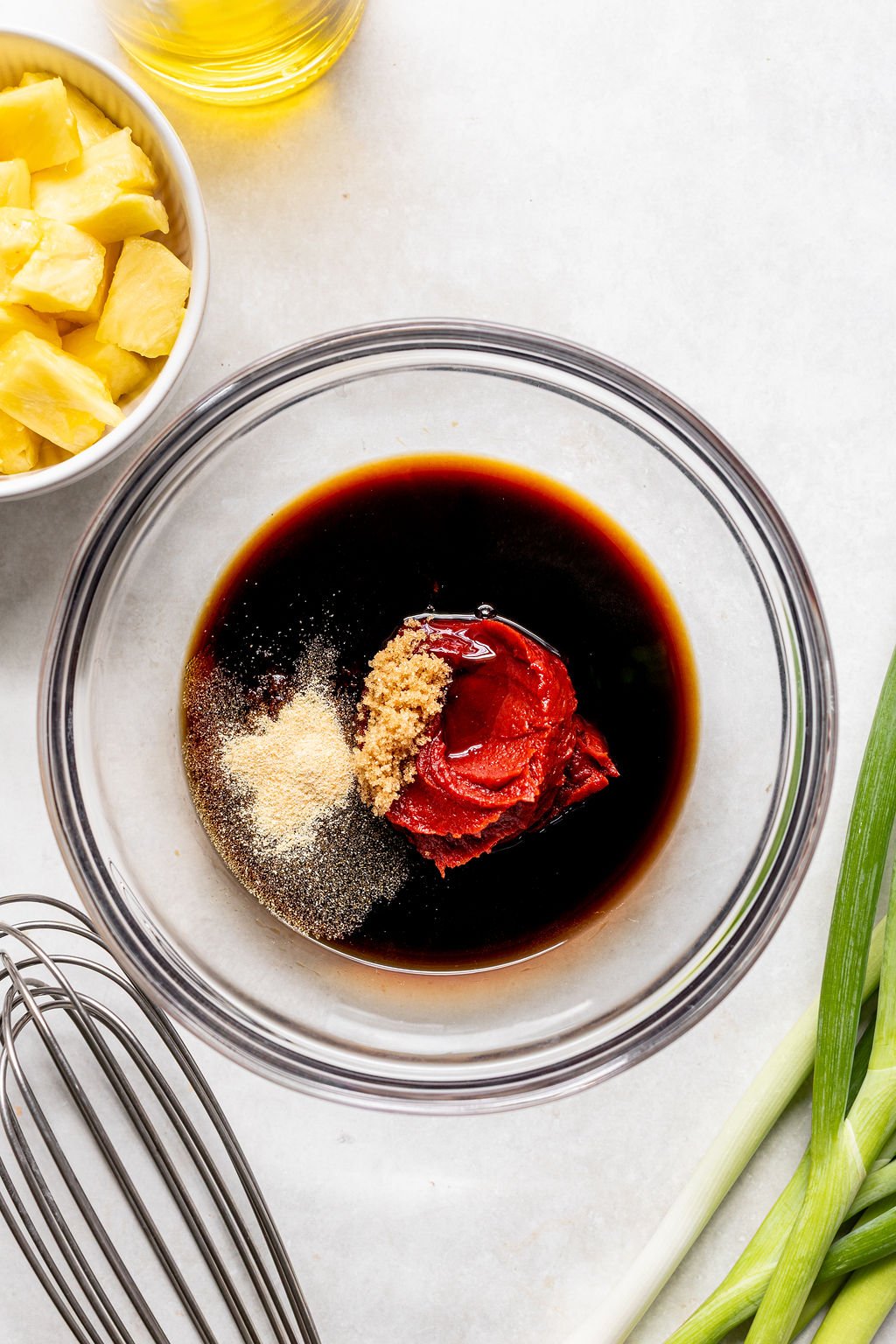 A glass bowl containing soy sauce, tomato paste, brown sugar, and seasonings, next to a whisk, chopped pineapple in a bowl, green onions, and a bottle of oil.