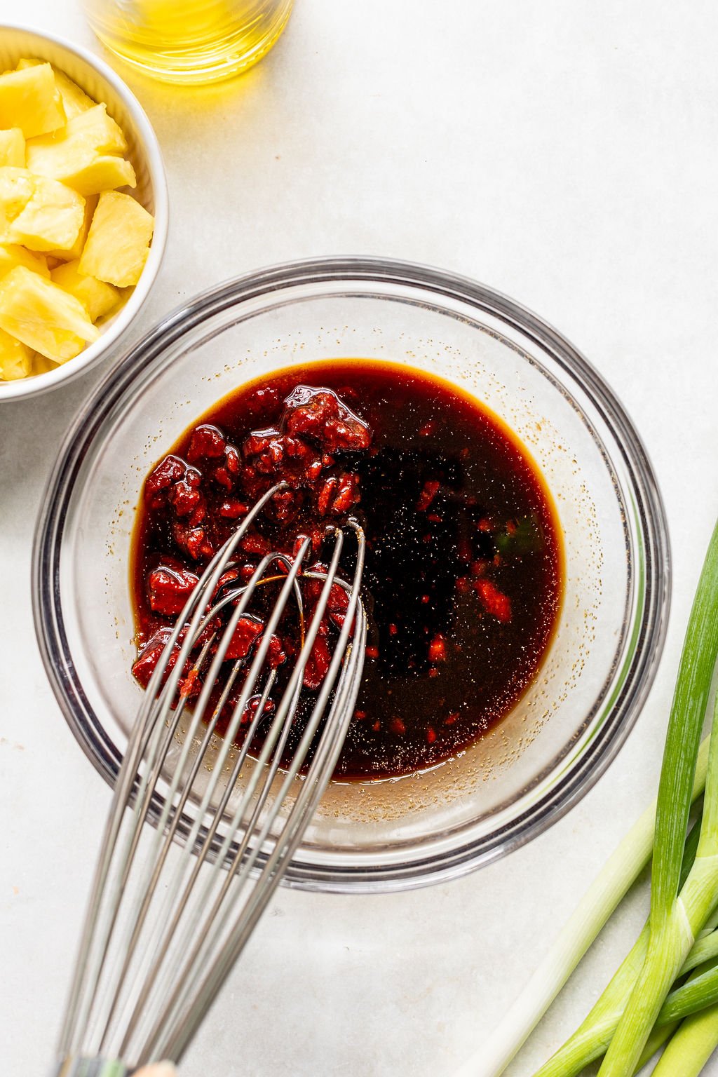 A whisk mixing a dark sauce with chopped ingredients in a glass bowl, next to a bowl of pineapple chunks and green onions on a white surface.