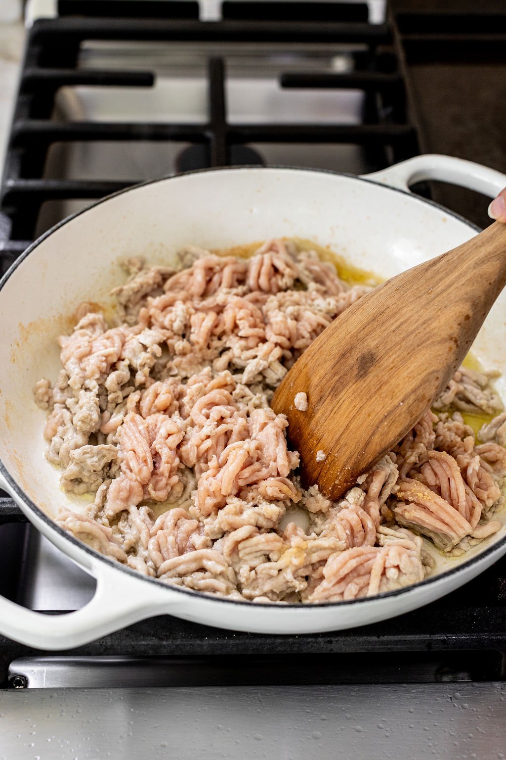 Ground meat is being cooked and stirred with a wooden spatula in a white skillet on a stovetop.