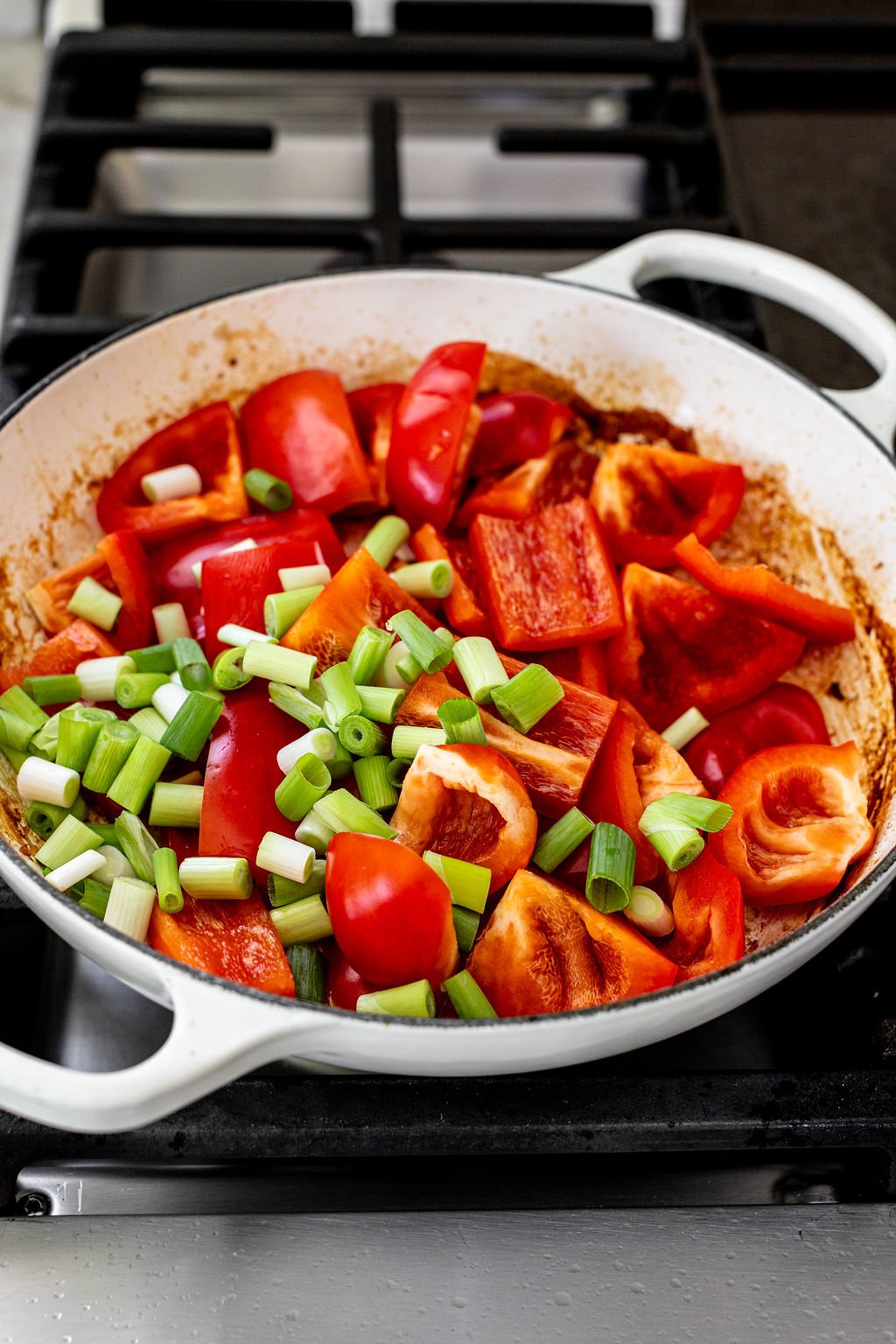 A white pan on a stove contains chopped red bell peppers and sliced green onions, ready to be cooked.
