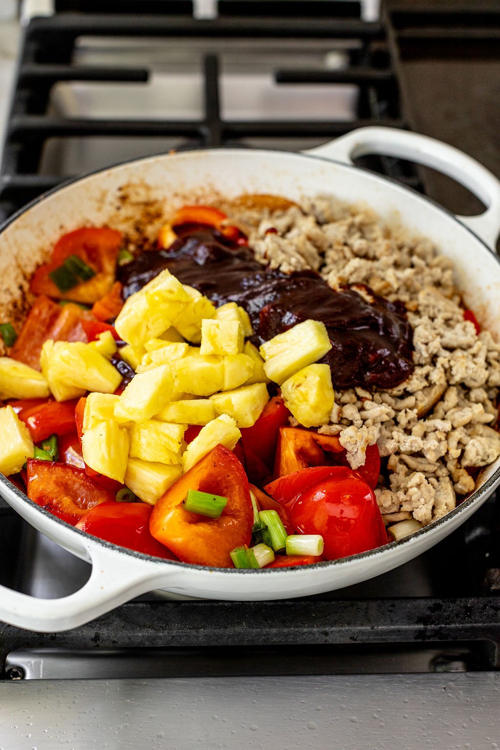 A white pot on a stove contains chopped tomatoes, pineapple chunks, ground meat, green onions, and a dark sauce, ready to be mixed and cooked.