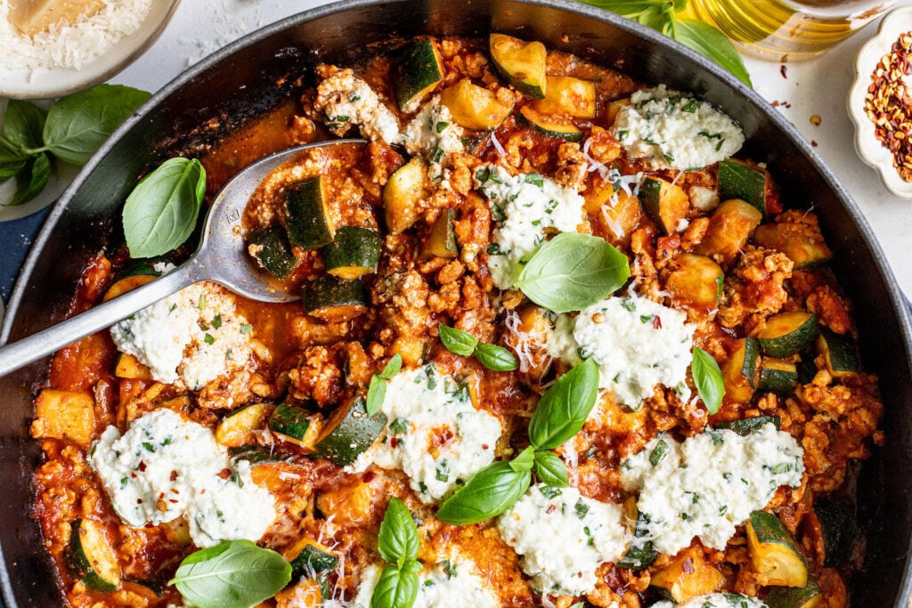 A skillet filled with a tomato-based dish containing ground meat, zucchini, dollops of ricotta cheese, and fresh basil leaves, with a serving spoon on the side.