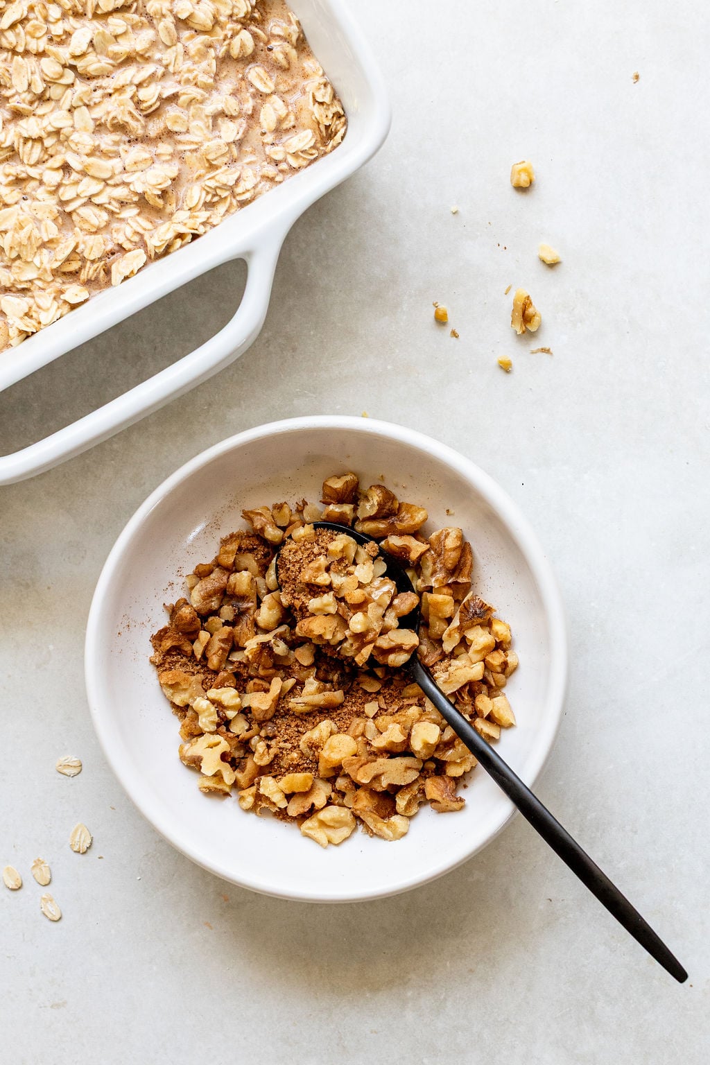 A white bowl with chopped walnuts and a black spoon, next to a white baking dish filled with an oat mixture on a light countertop.