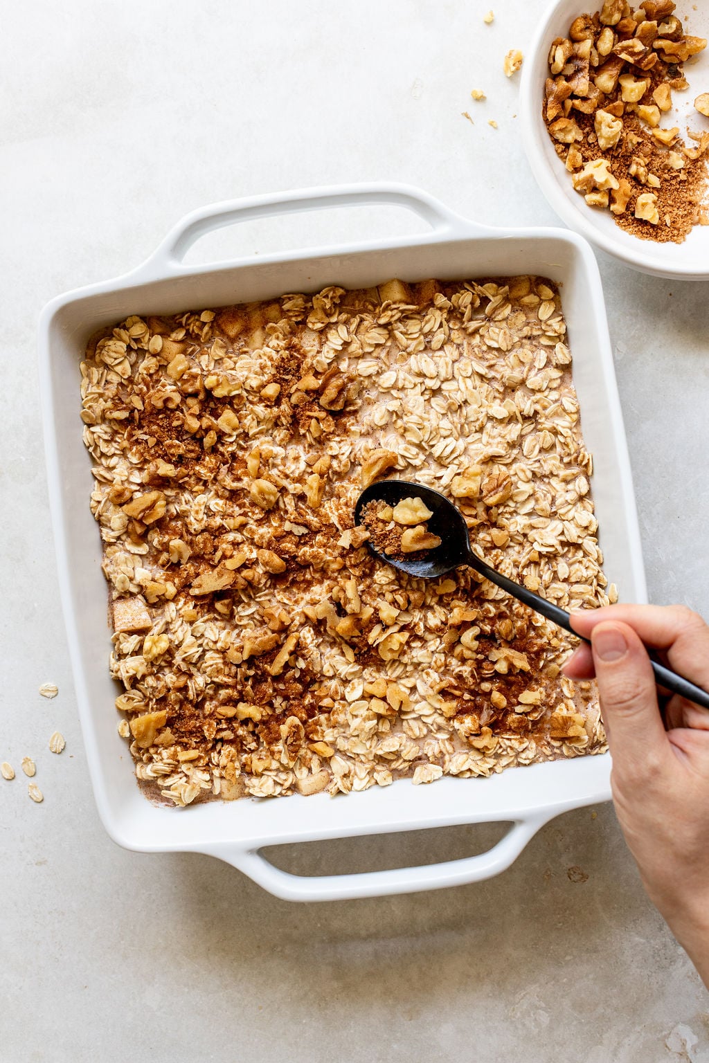 A hand sprinkles chopped walnuts and cinnamon over a square baking dish filled with oats, with a bowl of walnuts and cinnamon nearby.