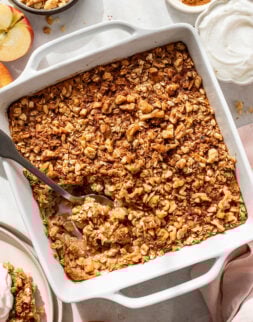 A square baking dish filled with baked apple crisp topped with oats and chopped nuts, with a serving spoon inside.